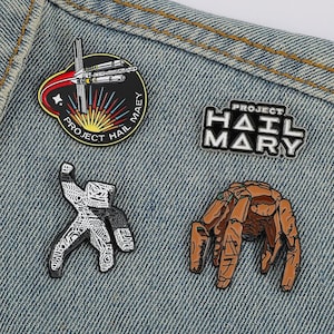 May include: Four enamel pins featuring Project Hail Mary themes: a circular mission patch with a spacecraft, a black and white text logo reading Project Hail Mary, a white and black patterned figure, and a brown, multi-legged creature design.