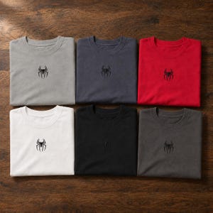 Op de afbeelding: Zes T-shirts met ronde hals gerangschikt in twee rijen, voorzien van een klein zwart geborduurd spinnenlogo in het midden op de borst. De shirts zijn verkrijgbaar in lichtgrijs, donkergrijs, rood, wit, zwart en antraciet, allemaal met korte mouwen.