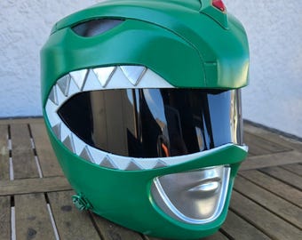 May include: A green helmet featuring a dark tinted visor, a silver mouthplate, and a row of silver triangular teeth accents along the visor edge. A small red diamond-shaped detail is positioned at the top center of the helmet shell.