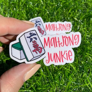 May include: Two white stickers with red and black text. One sticker features a cartoon illustration of a mahjong tile with the Chinese characters "East Wind" in red. The other sticker says "Mahjong Junkie".