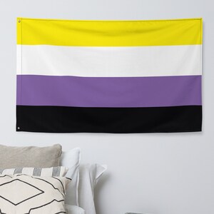 Könnte beinhalten: Eine Non-Binary-Pride-Flagge mit horizontalen Streifen in Gelb, Weiß, Lila und Schwarz. Die Flagge hängt an einer weißen Wand über einem Bett mit Kissen und einem hölzernen Beistelltisch.