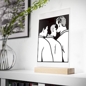 Könnte beinhalten: Schwarzweiß-Illustration von drei Personen, die eng beieinander stehen. Die Person in der Mitte hat einen kahlen Kopf mit bunten Punkten. Die Illustration ist auf einer durchsichtigen Acrylplatte mit einem Holzsockel gedruckt.