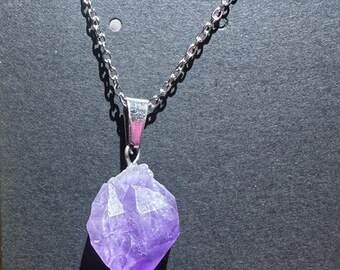 May include: A silver chain necklace with a faceted, raw amethyst pendant. The purple crystal is suspended from a silver bail and chain, set against a dark background. The necklace is a piece of jewelry.