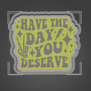 May include: A yellow sticker with a gray outline and the text "HAVE THE DAY YOU DESERVE" in a retro font. A stylized skeleton hand with two fingers raised in a peace sign is to the left of the text.