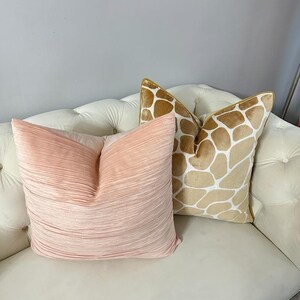 May include: Two decorative throw pillows on a white couch. One pillow is a light pink with a pleated texture. The other pillow is a tan and white patterned pillow with a giraffe print.