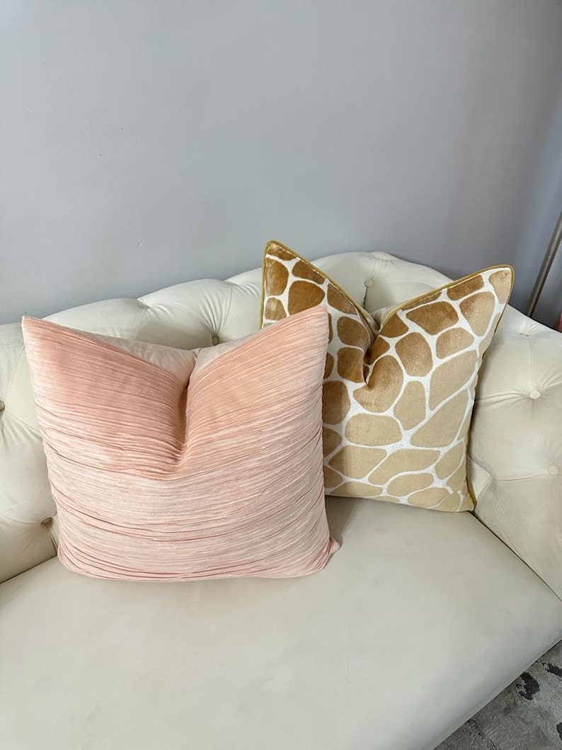 May include: Two decorative throw pillows on a white couch. One pillow is a light pink with a pleated texture. The other pillow is a tan and white patterned pillow with a giraffe print.