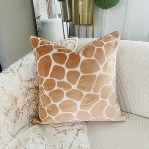 May include: A tan velvet throw pillow with a brown giraffe print pattern. The pillow is on a white couch with a gold and white patterned throw blanket.