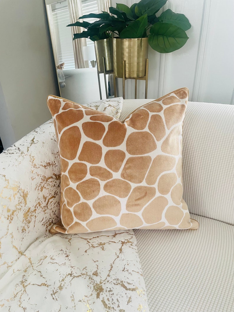 May include: A tan velvet throw pillow with a brown giraffe print pattern. The pillow is on a white couch with a gold and white patterned throw blanket.