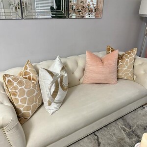 May include: A white tufted sofa with four throw pillows. The pillows are in various patterns and colors, including beige with brown spots, white with silver swirls, and a solid light pink. The sofa is in a living room setting with a glass coffee table and a rug.