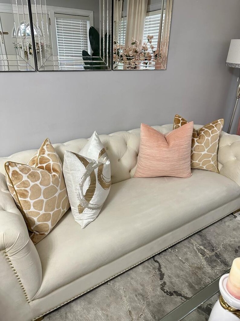 May include: A white tufted sofa with four throw pillows. The pillows are in various patterns and colors, including beige with brown spots, white with silver swirls, and a solid light pink. The sofa is in a living room setting with a glass coffee table and a rug.