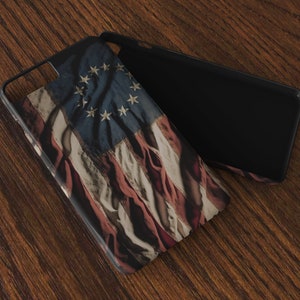 May include: A black phone case with a distressed American flag design featuring 13 stars. The case is lying on a wooden surface.