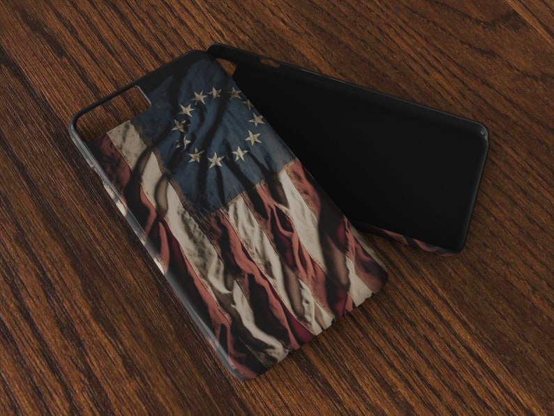 May include: A black phone case with a distressed American flag design featuring 13 stars. The case is lying on a wooden surface.