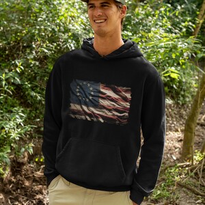 May include: A black hoodie with a distressed American flag design featuring 13 stars in a circle. The flag is faded and has a vintage look.