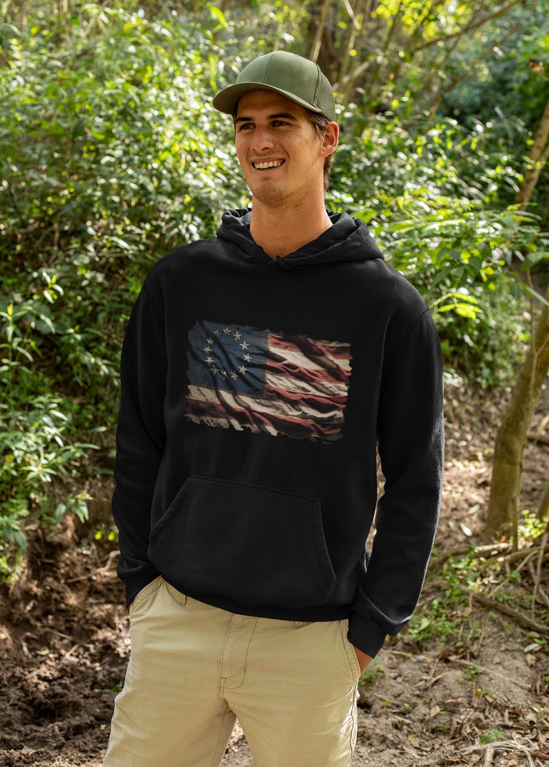 May include: A black hoodie with a distressed American flag design featuring 13 stars in a circle. The flag is faded and has a vintage look.