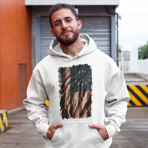 May include: A white hooded sweatshirt with a vintage-style print of the Betsy Ross flag. The flag is faded and distressed, with a red, white, and blue color scheme. The print is centered on the sweatshirt.