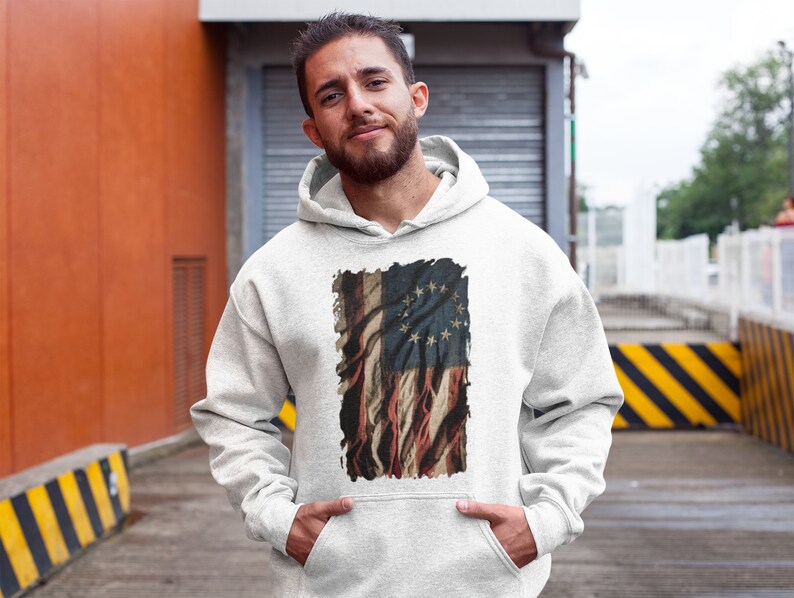 May include: A white hooded sweatshirt with a vintage-style print of the Betsy Ross flag. The flag is faded and distressed, with a red, white, and blue color scheme. The print is centered on the sweatshirt.