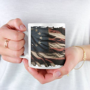 May include: White ceramic mug with a distressed American flag design. The flag has 13 stripes and 13 stars.