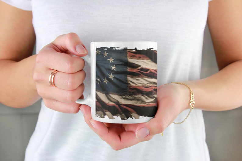 May include: White ceramic mug with a distressed American flag design. The flag has 13 stripes and 13 stars.