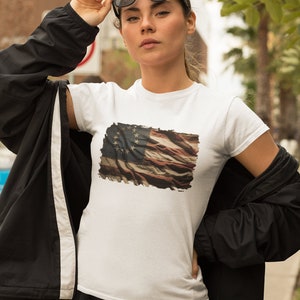 May include: A white t-shirt with a distressed American flag print featuring 13 stars. The flag is faded and has a vintage look. The model is wearing a black jacket and sunglasses.