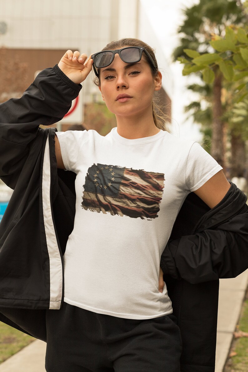 May include: A white t-shirt with a distressed American flag print featuring 13 stars. The flag is faded and has a vintage look. The model is wearing a black jacket and sunglasses.