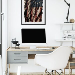 May include: A white desk with a wooden top and a gray drawer. A white chair with wooden legs is pulled back from the desk. A computer monitor is on the desk, along with a black lamp and a black bowl with writing utensils. A framed print of a distressed American flag with 13 stars hangs on the wall above the desk.