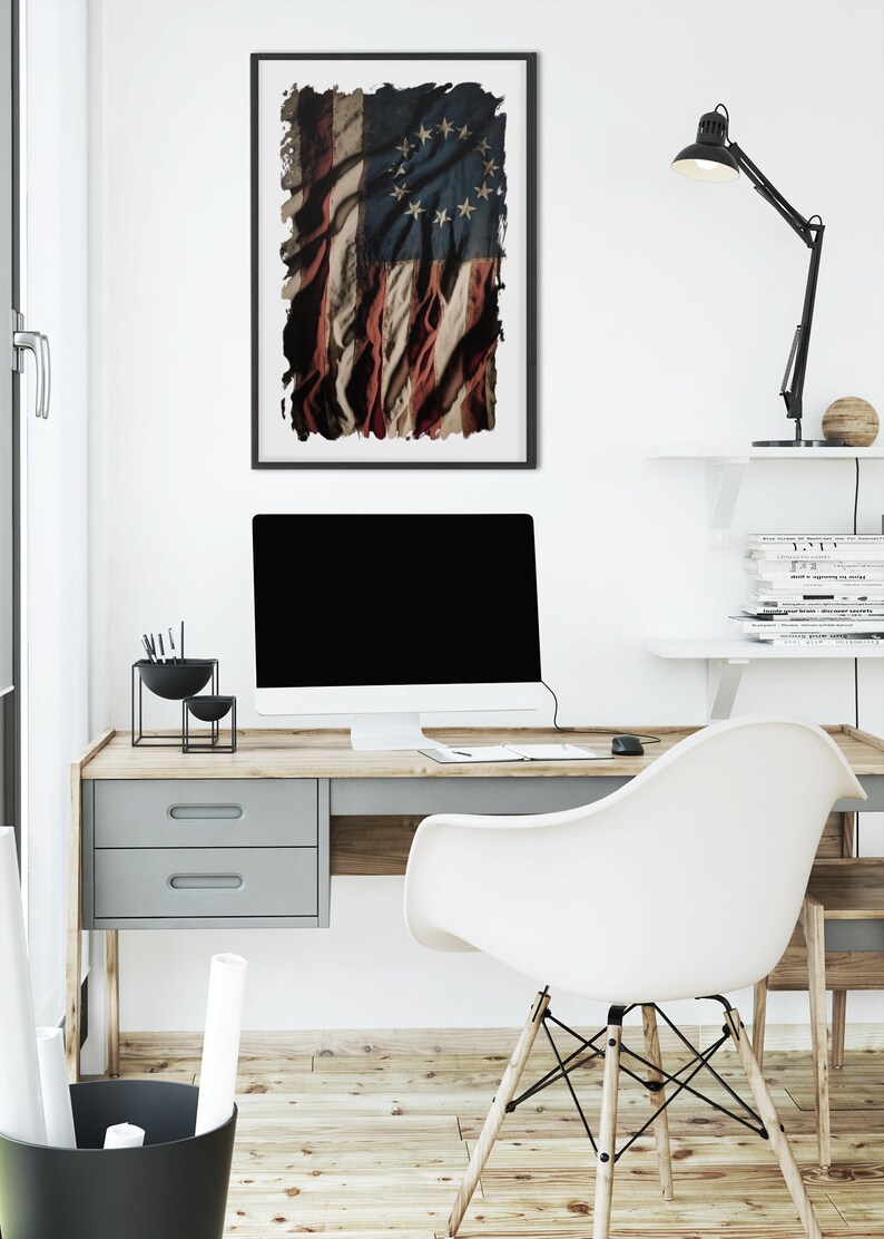 May include: A white desk with a wooden top and a gray drawer. A white chair with wooden legs is pulled back from the desk. A computer monitor is on the desk, along with a black lamp and a black bowl with writing utensils. A framed print of a distressed American flag with 13 stars hangs on the wall above the desk.
