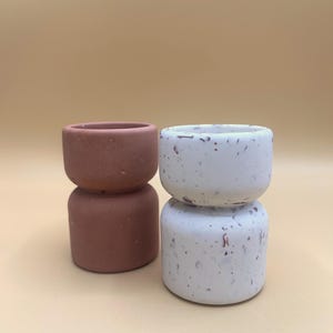 Peut inclure: Deux petits pots de fleurs empilés. L'un est de couleur brun rougeâtre uni, et l'autre est blanc avec des accents bruns mouchetés. Les pots ont une forme arrondie et sont faits d'un matériau texturé.
