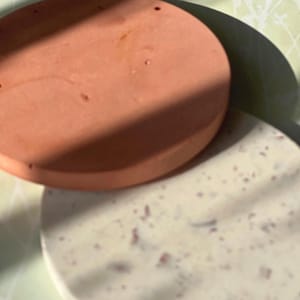 Peut inclure: Deux dessous de verre ronds. L'un est de couleur terre cuite unie, et l'autre est blanc cassé moucheté avec des accents rouges. Les dessous de verre sont empilés, avec le dessous de verre en terre cuite sur le dessus. Le fond est vert clair avec un motif floral.