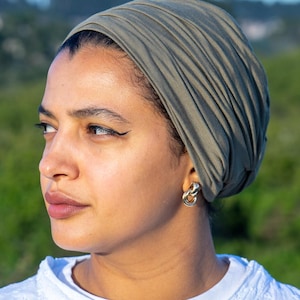 May include: An olive green head wrap is worn by a person. The head wrap is made of a soft, draped fabric. Gold-colored chain link earrings are worn. The person is wearing a white, embroidered shirt. The background is a blurred green field and blue sky.
