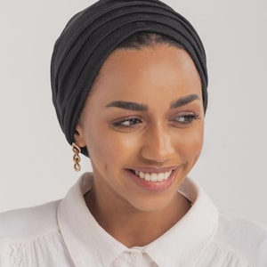 May include: A black turban-style head covering made from a ribbed fabric. The person is wearing gold earrings and a white shirt.