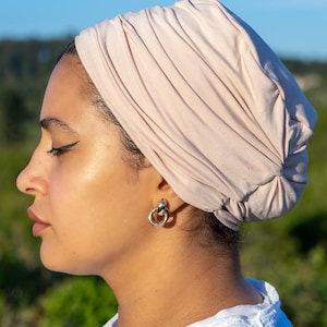 Könnte beinhalten: Ein hellbeige Turban-Kopfbedeckung. Der Stoff ist gerafft und drapiert, wodurch ein strukturiertes Aussehen entsteht. Die Person trägt silberne Creolen und ein weißes Oberteil. Der Hintergrund zeigt grünes Laub und einen blauen Himmel.