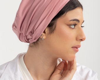 May include: A dusty rose colored turban style head wrap made of soft fabric. The head wrap is worn by a person with dark hair.