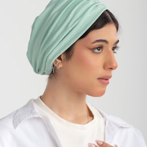 May include: A light green turban made of soft fabric. The turban is folded and draped over the head, creating a stylish and comfortable look.