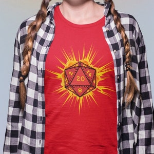 Op de afbeelding: Een rood t-shirt met een zonnesterontwerp en een twintigzijdige dobbelsteen in het midden. De dobbelsteen toont de nummers 20, 8 en 2. Het t-shirt wordt gedragen met een zwart-wit geruit overhemd.