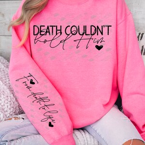 May include: A bright pink sweatshirt with the text "DEATH COULDN'T hold Him" in black script. The sleeve features the text "From death to life" in a vertical script with black hearts. A casual, comfortable top.