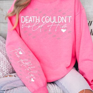 May include: Bright pink sweatshirt with the text "Death Couldn't Hold Him" in white script. The sleeve features the text "From death to life" in white script. Small gray heart and kiss-shaped patterns are visible.