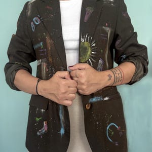 Könnte beinhalten: Ein dunkelbrauner Blazer mit handgemalten Motiven, darunter der Schriftzug 80s, ein Mikrofon, eine Sonne und Rollschuhe. Die Jacke hat lange Ärmel, ein steigendes Revers, einen Ein-Knopf-Verschluss und zwei Vordertaschen mit Klappen.