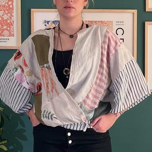 Op de afbeelding: Een patchwork wikkel top met een mix van patronen en kleuren. De top heeft strepen, bloemenprints en effen stukken in wit, roze, groen en olijf. De mouwen zijn wijd en de top heeft een wikkel sluiting.