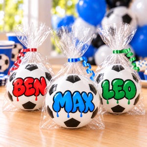 May include: Three soccer balls individually wrapped in clear plastic bags, each tied with a curly ribbon. The balls feature black and white panels with personalized names in colorful, dripping-style font: Ben in red, Max in blue, and Leo in green.