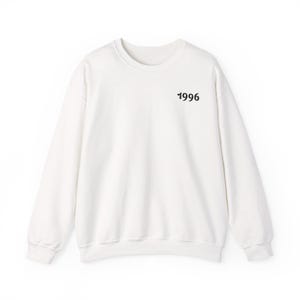 Puede incluir: Una sudadera blanca de manga larga con cuello redondo que presenta el número 1996 impreso en una fuente negra, estilizada y redondeada en la parte superior izquierda del pecho. La prenda tiene cuello, puños y dobladillo acanalados.