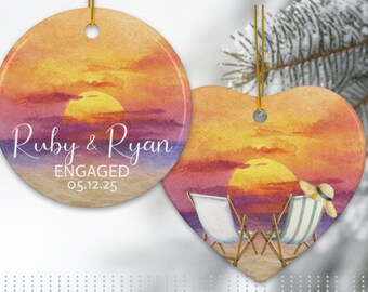 May include: Two ceramic ornaments with a sunset design. One is round with the names "Ruby & Ryan" and the word "ENGAGED" with a date. The other is heart-shaped with beach chairs and a hat. Both have gold hanging strings.