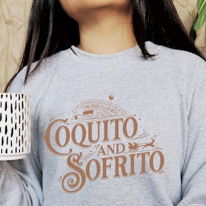 Puede incluir: Una sudadera gris con las palabras "Coquito and Sofrito" en escritura marrón. La sudadera también presenta un gráfico de un timón y un trineo con renos. En la imagen se sostiene una taza blanca con guiones negros.