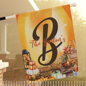 Peut inclure: Un drapeau de jardin rectangulaire arborant une grande lettre B marron foncé avec le texte The Benton's en écriture cursive. Le fond présente une illustration à l'aquarelle de citrouilles orange, une lanterne, un panier et des bottes de foin.