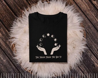 May include: Black t-shirt featuring a graphic of the moon phases held between two hands. The text "The Moon Made Me Do It" is printed below the graphic. The shirt is folded and placed on a white fluffy surface.