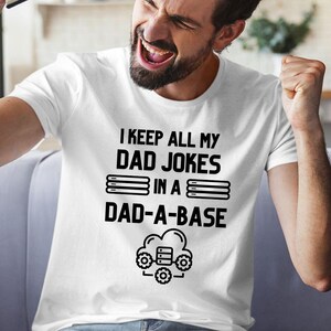 May include: White t-shirt with the text "I KEEP ALL MY DAD JOKES IN A DAD-A-BASE" in black. The design includes stacked lines and a cloud with gears. The person wearing the shirt is smiling and has their fist raised.