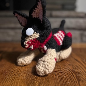 May include: A handmade crochet dog toy in black, red, and beige. The dog has large ears, white button eyes, and a red and white striped collar. The toy is on a wooden surface.