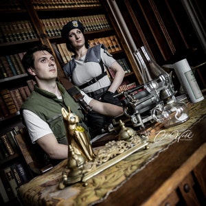 May include: Two people dressed in costumes stand in a room with a wooden bookcase and a table. The person on the left is wearing a green shirt and a vest. The person on the right is wearing a black hat and a gray shirt with a black vest. The table has a gold decorative object on it.
