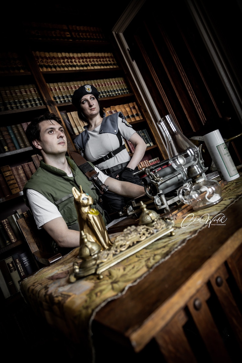 May include: Two people dressed in costumes stand in a room with a wooden bookcase and a table. The person on the left is wearing a green shirt and a vest. The person on the right is wearing a black hat and a gray shirt with a black vest. The table has a gold decorative object on it.
