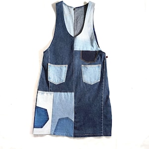 Op de afbeelding: Een mouwloze spijkerjurk met een patchwork ontwerp. De jurk heeft een V-hals en twee voorzakken. De denim is een mix van lichte en donkere blauwtinten, wat een unieke en stijlvolle look creëert.