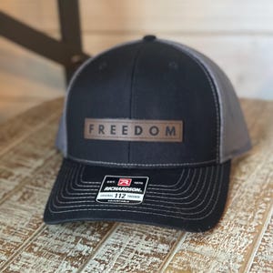 May include: Black and gray trucker hat with the word "FREEDOM" on a brown leather patch. The hat has a curved brim and an adjustable strap. The hat is sitting on a wooden surface.